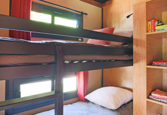 Glampingværelse med køjesenge, træreoler, opbevaringshylder og vinduer med røde gardiner i dagslys.