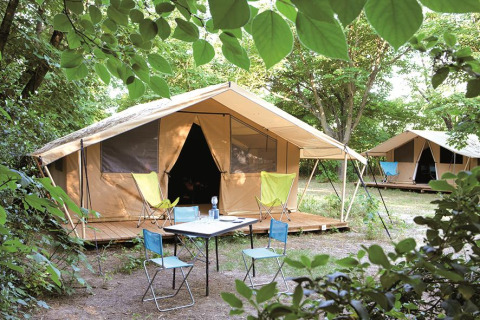 Safari Toile&bois tent Classic in nature, with a wooden deck, camping chairs and table among trees.