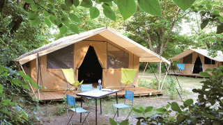 Safari Toile&bois tent Classic in de natuur met houten terras, campingstoelen en tafel tussen bomen.