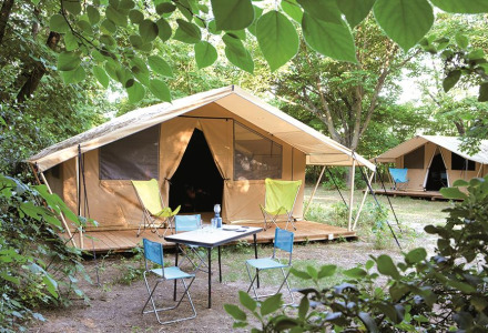 Safari Toile&bois tent Classic in de natuur, met houten terras, campingstoelen en tafel onder bomen.