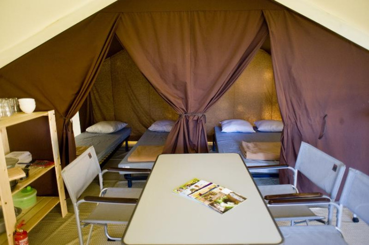 Interior of a safari tent featuring four single beds, a dining table, chairs, and shelves for glamping.