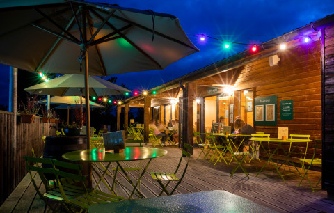 Udendørs caféområde ved Huttopia Etang de Fouché i Bourgogne, Frankrig med farverige lyskæder om aftenen.