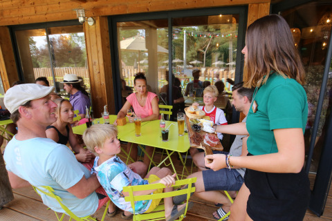 Una familia disfruta al aire libre en Huttopia Etang de Fouché, Francia, mientras una camarera sirve comida.