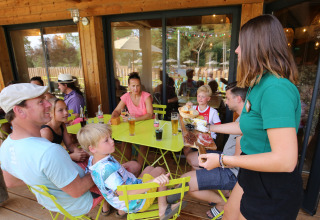 En familie sidder ved et bord udendørs på Huttopia Etang de Fouché, mens en tjener serverer mad.