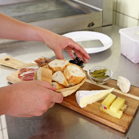 Iemand schikt een plateau met brood, kaas, charcuterie, olijven en augurken op een keukentafel.