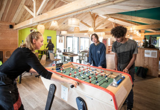 Tres jóvenes adultos juegan futbolín en una acogedora sala común de madera en Huttopia Etang de Fouché, Francia.