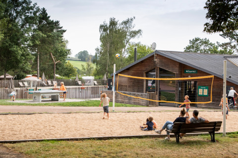 Niños juegan al voleibol en una cancha de arena en un parque vacacional, mientras adultos observan en Francia.