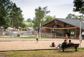 Kinderen spelen volleybal op het zand bij een vakantiepark, volwassenen kijken toe vanaf een bankje.