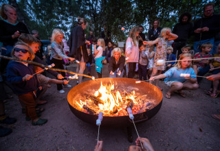 Children and adults roasting marshmallows over a campfire at Huttopia Etang de Fouché holiday park in France.