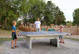 Two children play outdoor table tennis at a holiday park while an adult supervises, surrounded by greenery.