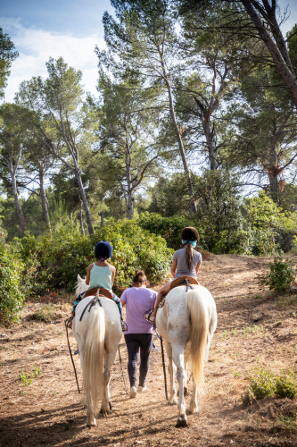 Twee kinderen rijden op witte paarden door een bos bij Fontvieille, begeleid door een volwassene te voet.