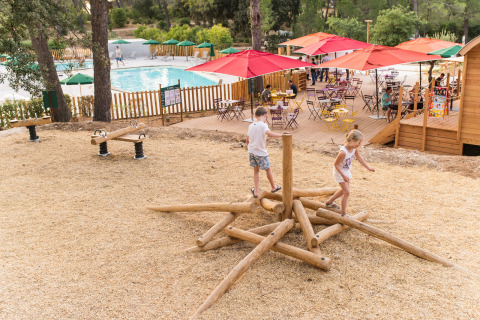 Kinderen spelen op houten speeltoestel bij zwembad en terras in Huttopia Fontvieille, Frankrijk.