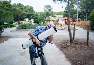 A man in a checkered shirt uses a telescope at a holiday park surrounded by trees in Provence, France.