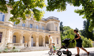 Familia pasea con carrito y bicicleta infantil bajo árboles frente a un edificio histórico en Fontvieille, Provenza.