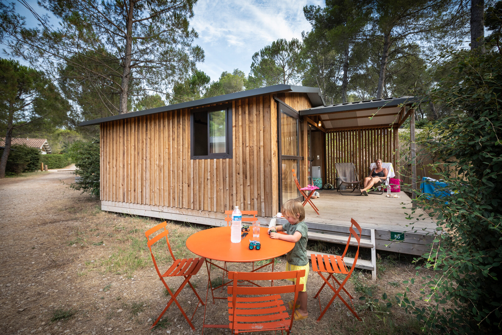 Een kind speelt aan een oranje tafel voor een houten lodge bij Chalet Evasion, Huttopia Fontvieille, Frankrijk.