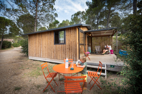 Un niño juega en una mesa naranja frente a una cabaña de madera en Chalet Evasion, Huttopia Fontvieille, Francia.