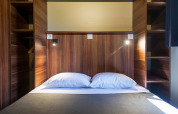 Cama doble moderna en chalet de madera glamping con estantes integrados y luces de lectura a ambos lados.