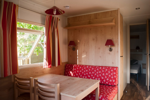 Interior acogedor de cabaña con muebles de madera, banco rojo y ventana abierta en Huttopia Fontvieille.