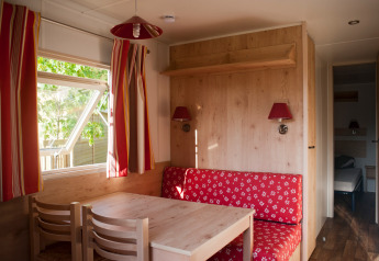 Interior acogedor de cabaña con muebles de madera, banco rojo y ventana abierta en Huttopia Fontvieille.