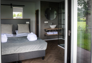 Habitación de lodge moderna con cama doble, baño abierto y gran ventanal con vistas verdes en Países Bajos.