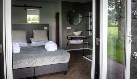 Modern lodge room with double bed, open bathroom, large windows, and green outdoor view in the Netherlands.