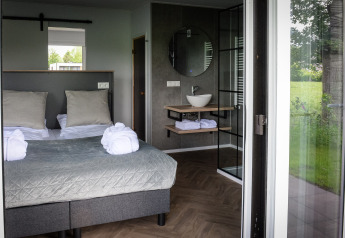 Modern lodge room with double bed, open bathroom, large windows, and green outdoor view in the Netherlands.