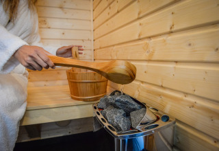 Persona in accappatoio versa acqua sulle pietre calde della sauna al Panorama Wellness Lodge, Vakantiepark Hölte.