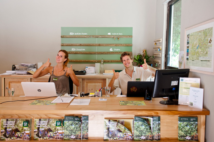 Receptionists giving thumbs up and smiling at the front desk of Huttopia Gorges du Verdon holiday park, France.