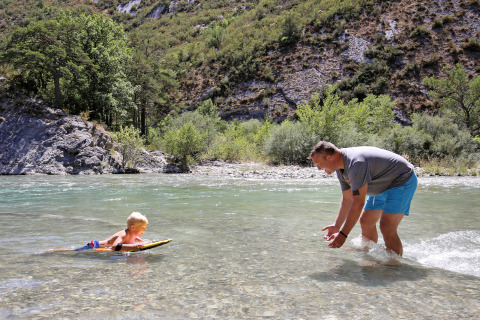 Un hombre y un niño juegan en un río poco profundo en Huttopia Gorges du Verdon con colinas de fondo.