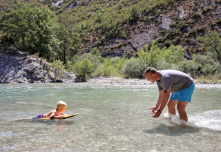 A man and child play in the shallow river at Huttopia Gorges du Verdon holiday park with hills behind them.