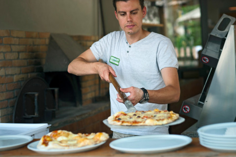 Hombre cortando pizza en cocina con horno de leña y platos, en Huttopia Gorges du Verdon, Hauts-de-France.