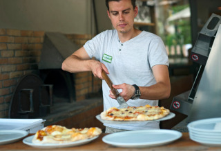 Mand skærer pizza i et køkken med brændeovn, med flere tallerkener på bordet, på Huttopia Gorges du Verdon.