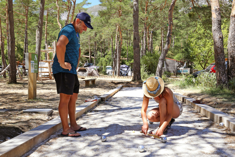 Twee mensen spelen jeu de boules op een grindbaan bij Huttopia Gorges du Verdon, omgeven door bos.