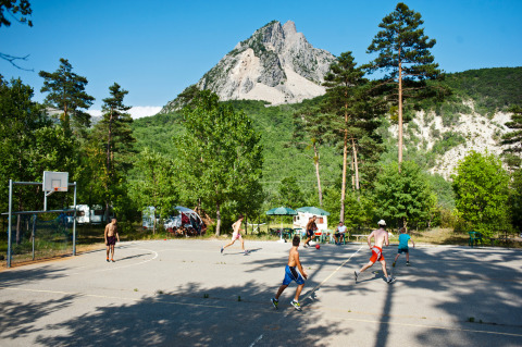 Des enfants jouent au basket sur un terrain extérieur entouré d’arbres et montagnes à Huttopia Gorges du Verdon.