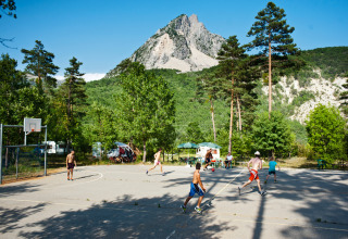Niños jugando baloncesto en una cancha al aire libre rodeada de árboles y montañas en Huttopia Gorges du Verdon.