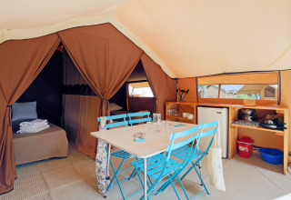 Interieur van een klassieke bungalow-safaristent met eettafel, blauwe stoelen, bed en keukentje.