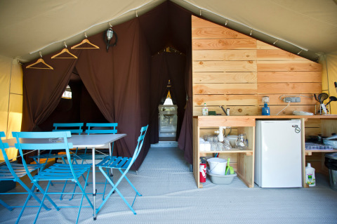 Interno di una tenda safari con tavolo e sedie blu, angolo cottura, mini frigo e tende marroni.