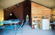Interior of a safari tent with blue dining set, kitchenette, mini fridge, brown curtains, and hangers.
