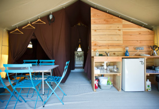 Interno di una tenda safari con tavolo e sedie blu, angolo cottura, mini frigo e tende marroni.