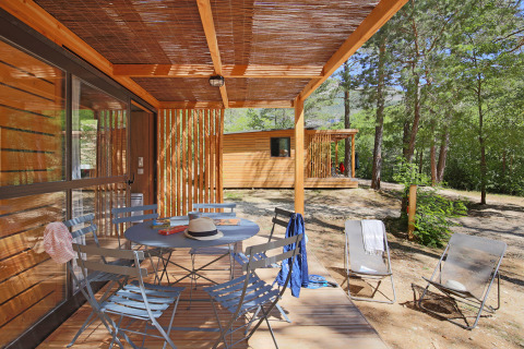 Træveranda med borde og stole på Chalet Evasion ved Huttopia Gorges du Verdon, omgivet af skov i Frankrig.