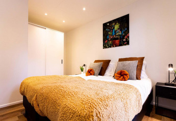 Chambre moderne dans un lodge avec lit double, couverture dorée, coussins et tableau mural coloré.