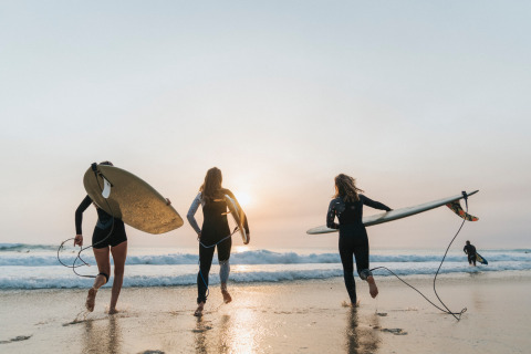 Feriegæster med surfbrætter løber ud i havet under solnedgang ved Huttopia Lac de Carcans i Frankrig.