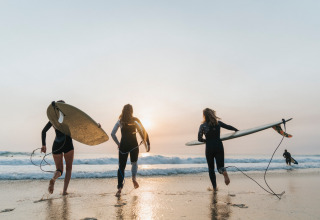 Feriegæster med surfbrætter løber ud i havet under solnedgang ved Huttopia Lac de Carcans i Frankrig.