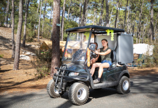 Two young people drive a golf cart through a wooded campsite at Huttopia Lac de Carcans holiday park, France.