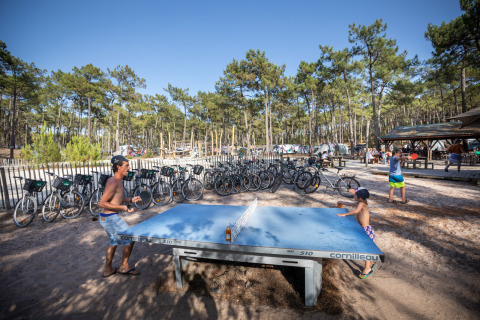Parque vacacional en Huttopia Lac de Carcans, Francia, con gente jugando al ping-pong y bicicletas cercanas.