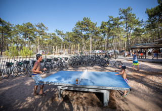 Feriepark scene ved Huttopia Lac de Carcans, Frankrig, med folk der spiller bordtennis og cykler i baggrunden.