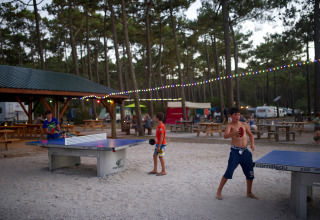 Børn spiller bordtennis udendørs under lyskæder ved feriestedet Huttopia Lac de Carcans i Frankrig.