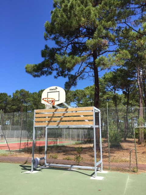 Canasta de baloncesto al aire libre en Huttopia Lac de Carcans, rodeada de pinos y cielo azul despejado.