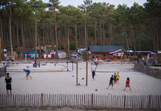Børn og voksne spiller volleyball i sandet, omgivet af skov og telte i Huttopia Lac de Carcans, Frankrig.