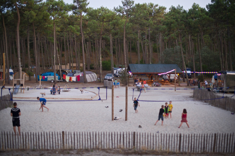 Kinder und Erwachsene spielen Volleyball im Sand in Huttopia Lac de Carcans, umgeben von Wald und Zelten.